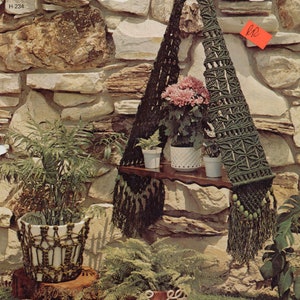 May include: A green macrame plant hanger with wooden shelf and tassels. The hanger is hanging from a stone wall and has three potted plants on the shelf. The image includes the text "MACRAME MAGIC" and "H-234".