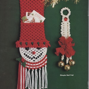 May include: A red and white crocheted snowman card holder with a pocket for cards and a white crocheted bell pull with a red bow and gold bells. The snowman has a red nose, black eyes, and a green holly sprig.