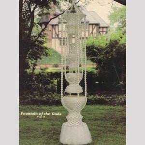 May include: A white macrame hanging planter with three tiers, labeled "Fountain of the Gods, Page 13". The planter is hanging in a garden setting.