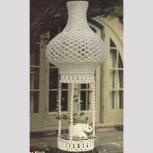 Op de afbeelding: Witte macramé plantenhanger met een rond schap en een klein wit olifantbeeldje. De hanger hangt aan een tak.