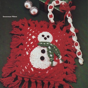 Puede incluir: Una almohada de muñeco de nieve de crochet rojo con detalles blancos y verdes. La almohada tiene un borde de flecos y tiene la forma de un muñeco de nieve con una bufanda verde y ojos negros. La almohada está sobre un fondo verde con un bastón de caramelo con cuentas y una caja de regalo.