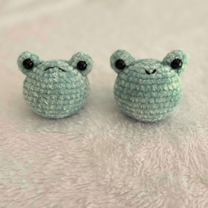 May include: Two small crocheted green frog plushies with black eyes. The frogs are sitting on a white fuzzy surface.