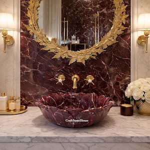 May include: A luxurious bathroom vanity with a burgundy floral-shaped sink. The vanity features a gold-framed mirror, gold fixtures, and sconces. A gold tray with bottles and a vase of white roses complete the elegant design.