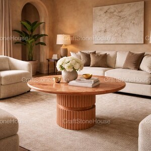 May include: A living room with a round, reddish-orange coffee table with a ribbed base. The table is in front of a cream sofa with brown cushions. A vase of white flowers sits on the table. The room has a neutral colour palette.