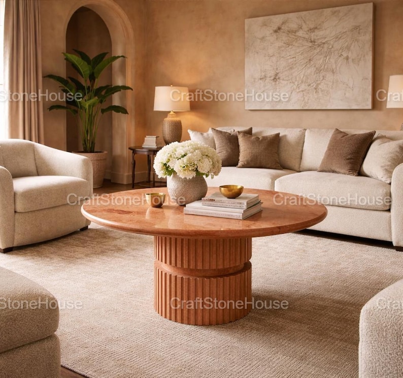 May include: A living room with a round, reddish-orange coffee table with a ribbed base. The table is in front of a cream sofa with brown cushions. A vase of white flowers sits on the table. The room has a neutral colour palette.