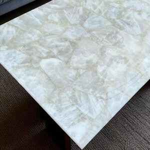 May include: A rectangular coffee table with a light-colored, marbled stone top. The stone has a translucent, cloudy appearance with subtle variations in color. The table's base is a dark, solid color, providing a contrast to the bright top.