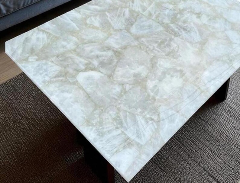 May include: A rectangular coffee table with a light-colored, marbled stone top. The stone has a translucent, cloudy appearance with subtle variations in color. The table's base is a dark, solid color, providing a contrast to the bright top.