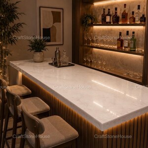 May include: A modern home bar with a white marble countertop and wooden accents. The bar features a built-in shelf with bottles and glasses, and is illuminated with warm lighting. Several bar stools are positioned in front of the bar.