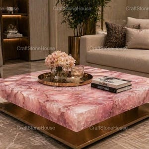 May include: A square coffee table with a rose quartz top and a gold-colored metal base. The table is decorated with a tray of flowers, candles, and two books. The table is in a living room setting.