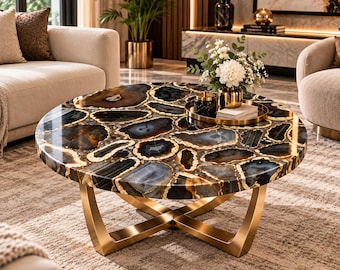 May include: A round coffee table featuring a tabletop composed of polished, multi-colored agate slices in shades of black, gray, and amber with gold-toned edges. The table is supported by a sturdy, intersecting gold-finished metal base.