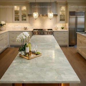 Peut inclure: Cuisine moderne avec un grand îlot central en marbre vert clair. La cuisine comprend des armoires blanches, des appareils électroménagers en acier inoxydable et un éclairage décoratif. Un plateau avec des bouteilles de vin, des verres et des fruits est posé sur l'îlot.
