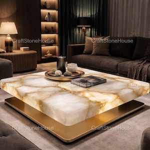 May include: A contemporary square coffee table featuring a glowing, translucent white and gold stone top. The table rests on a gold-coloured metal base. A tray with spheres and a book are on the table.