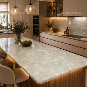 May include: A modern kitchen with a large, light-colored countertop island. The island has a wooden base and is illuminated with warm lighting. The kitchen features neutral-toned cabinets, a built-in oven, and pendant lights.