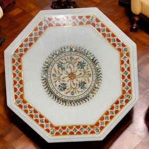 Puede incluir: Mesa octogonal de mármol blanco con intrincados incrustaciones florales. La mesa presenta un diseño circular central y un borde con detalles en rojo y dorado. Una pieza decorativa para salones.
