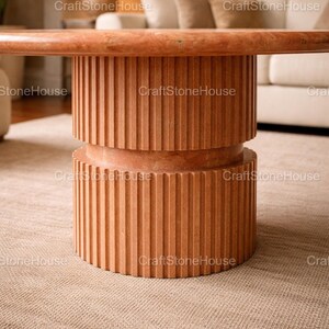 May include: A contemporary coffee table in a reddish-brown hue. The table has a cylindrical base with vertical grooved detailing. The tabletop is a matching colour. The table is set on a neutral-coloured rug.
