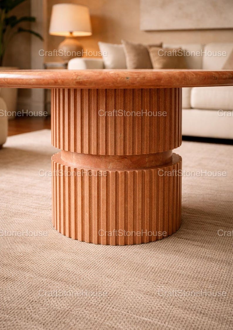 May include: A contemporary coffee table in a reddish-brown hue. The table has a cylindrical base with vertical grooved detailing. The tabletop is a matching colour. The table is set on a neutral-coloured rug.