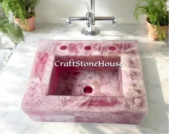Può includere: Un lavandino quadrato in quarzo rosa con un bacino rettangolare e tre fori per il rubinetto. Il lavandino è incassato in un piano di lavoro bianco con un rubinetto cromato. Il testo "CraftStoneHouse" è visibile sul lavandino.
