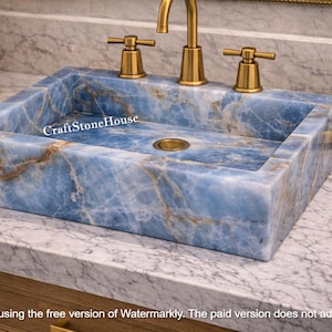 Può includere: Un lavabo rettangolare in marmo blu con rubinetti color oro. Il lavabo presenta un motivo venato naturale ed è posizionato su un piano di lavoro in marmo. Il rubinetto e le maniglie sono dorati. Il testo "CraftStoneHouse" è visibile.