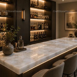 May include: A luxurious home bar with a marble countertop and built-in shelving. The shelves are stocked with bottles and glassware, and the bar is illuminated with warm lighting. There are bar stools and decorative vases on the counter.