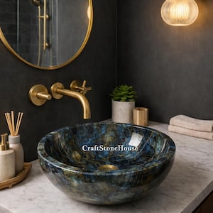 May include: A round, blue and gold stone vessel sink with a polished finish. The sink is set on a white marble countertop with gold fixtures and a round mirror. The sink is labeled "CraftStoneHouse".
