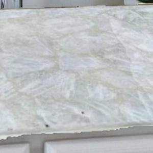 May include: A white countertop with a textured, marbled appearance. The surface has a light, translucent quality with subtle variations in color and pattern. The countertop is set on a white cabinet.