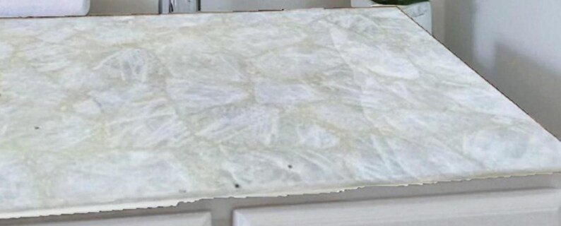 May include: A white countertop with a textured, marbled appearance. The surface has a light, translucent quality with subtle variations in color and pattern. The countertop is set on a white cabinet.