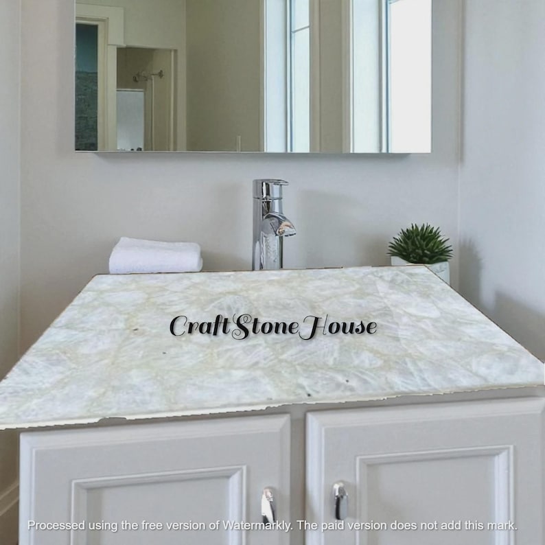 May include: A white marble countertop with a light pattern, installed over a white cabinet. A chrome faucet and a small potted plant are visible. The text "Craft Stone House" is displayed on the countertop.
