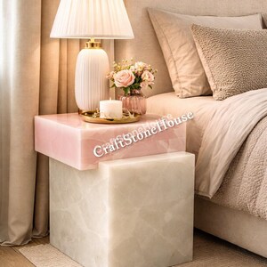 Könnte beinhalten: Ein modernes Schlafzimmer mit einem gestapelten Beistelltisch aus rosa und weißem Marmor. Eine weiße Lampe mit plissiertem Schirm, eine Vase mit rosa Rosen und eine Kerze stehen auf dem Tisch. Der Text "CraftStone House" ist sichtbar.