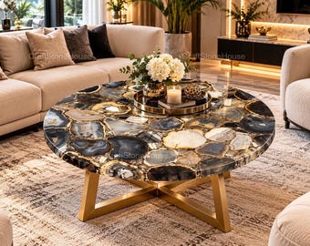 May include: A round coffee table featuring a tabletop made of polished, multi-colored agate slices in shades of black, gray, white, and gold. The table is supported by a gold-toned metal base with a cross-legged design, providing a stable foundation.