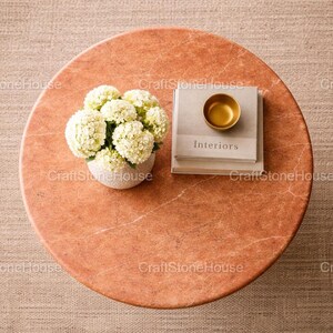 May include: A circular, reddish-orange marble coffee table with a white floral arrangement in a small, speckled vase. A stack of books with the word "Interiors" and a small gold bowl are on top of the books.