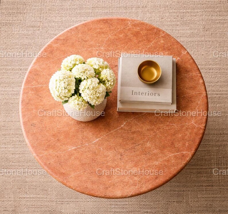 May include: A circular, reddish-orange marble coffee table with a white floral arrangement in a small, speckled vase. A stack of books with the word "Interiors" and a small gold bowl are on top of the books.