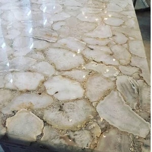 May include: A polished countertop with a unique design. The surface is composed of irregular, light-colored stone pieces with brown and beige edges, creating a mosaic-like pattern. The countertop reflects light, enhancing its glossy appearance.