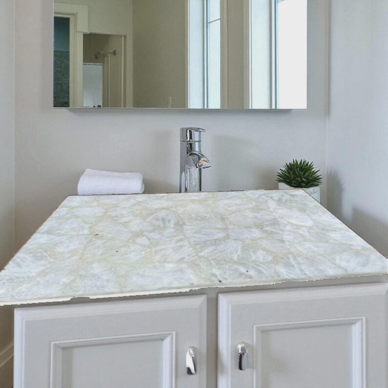 May include: A white bathroom vanity with a patterned countertop. The countertop has a mosaic design. A chrome faucet is mounted on the countertop. A small, potted plant and a rolled white towel are also on the countertop. The vanity has two doors with silver handles.
