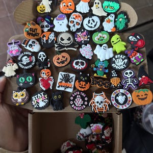 May include: A collection of Halloween-themed shoe charms, including ghosts, pumpkins, skulls, and other spooky characters. The charms are made of colorful silicone and feature various designs and sayings, such as "Stay Spooky" and "Save the Boobies".