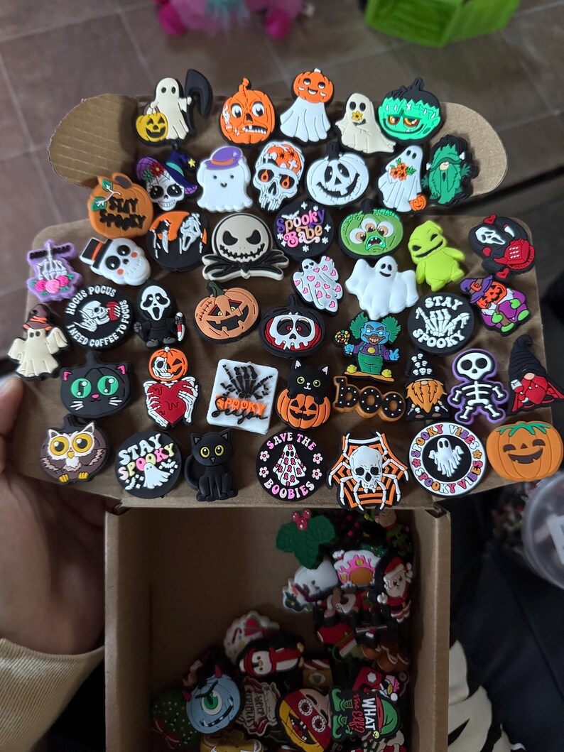 May include: A collection of Halloween-themed shoe charms, including ghosts, pumpkins, skulls, and other spooky characters. The charms are made of colorful silicone and feature various designs and sayings, such as "Stay Spooky" and "Save the Boobies".