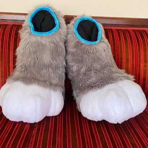 May include: A pair of furry gray and white paw-shaped costume feet. The feet have a white paw pad, gray fur, and a blue trim around the opening. The interior is black.