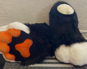 May include: A pair of black and white furry paw-shaped costume feet with orange paw pads. The feet are designed to resemble animal paws, with a plush, textured appearance. The interior of the foot is a quilted cream color.