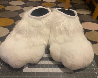 May include: A pair of white furry costume feet with black interiors. The plush feet are large and rounded, designed to resemble animal paws. The faux fur is long and fluffy, creating a soft, textured appearance. The feet are likely part of a larger costume.