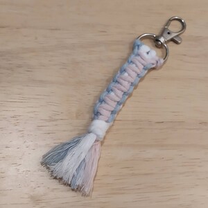 May include: A light blue, white and pink braided keyring with a silver key ring. The keyring has a tassel at the end.