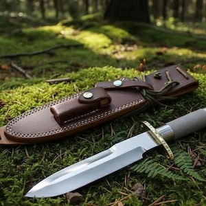 May include: A hunting knife with a silver blade and a textured gray handle, next to a brown leather sheath. The knife has a gold-colored guard and a gold-colored cap on the handle. The sheath has a button closure.