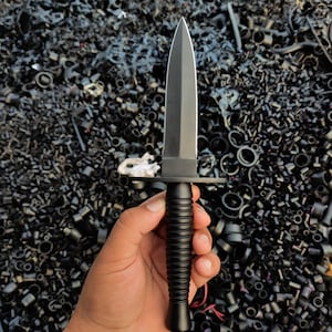 May include: A black dagger with a pointed blade and a ribbed handle. The dagger has a crossguard and is held in a hand. The background is a pile of black objects.
