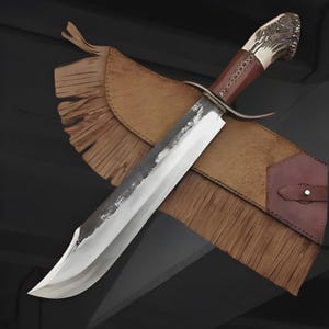 Könnte beinhalten: Ein großes, handgefertigtes Messer mit polierter Stahlklinge und braunem Ledergriff. Der Griff hat ein dekoratives Geweihdesign und wird mit einer Fransenlederscheide kombiniert. Das Messer ist etwa 38 cm lang.