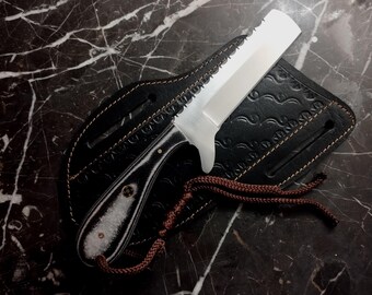 May include: A stainless steel knife with a serrated edge and a black and white handle rests in a black leather sheath. The sheath has a decorative embossed pattern and brown stitching. A brown cord is attached to the sheath.