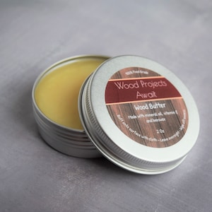May include: A small silver tin with a label that reads "Wood Projects Await Wood Butter Made with mineral oil, vitamin E and beeswax 2 oz Buff onto surface with cloth - Leave overnight". The tin is open and contains a yellow, waxy substance.