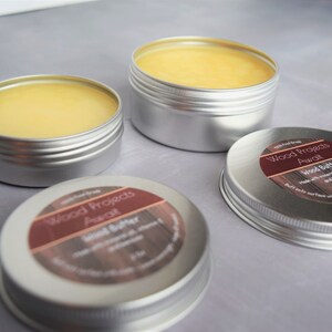 May include: Three silver tins of wood butter, labelled "Wood Projects Awali Wood Butter". The tins are filled with a yellow, waxy substance.