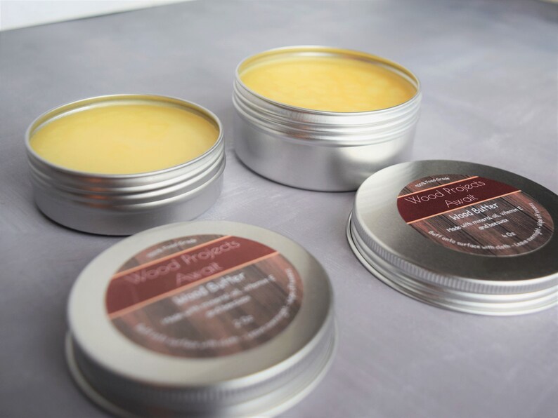 May include: Three silver tins of wood butter, labelled "Wood Projects Awali Wood Butter". The tins are filled with a yellow, waxy substance.