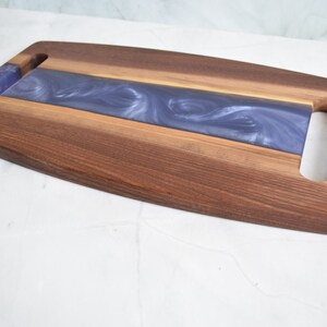 Op de afbeelding: Een houten serveerplank met een paarse harsinleg en twee uitgesneden handvatten. De plank is gemaakt van walnoot hout en heeft een natuurlijke afwerking.