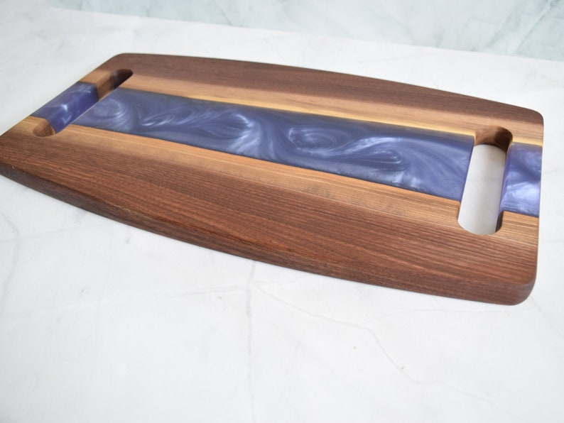 Op de afbeelding: Een houten serveerplank met een paarse harsinleg en twee uitgesneden handvatten. De plank is gemaakt van walnoot hout en heeft een natuurlijke afwerking.