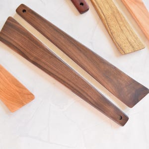 May include: A selection of wooden kitchen utensils, including two dark brown walnut spatulas with rounded edges and a hole in the handle. Other wooden utensils in various light brown shades are also visible.