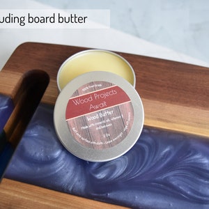 Op de afbeelding: Een klein blikje houtboter, met het label "Wood Projects Await", met de tekst "100% Food Grade" en "Made with mineral oil, vitamin E and beeswax". Het blikje staat op een houten snijplank met een blauwe harsinleg.